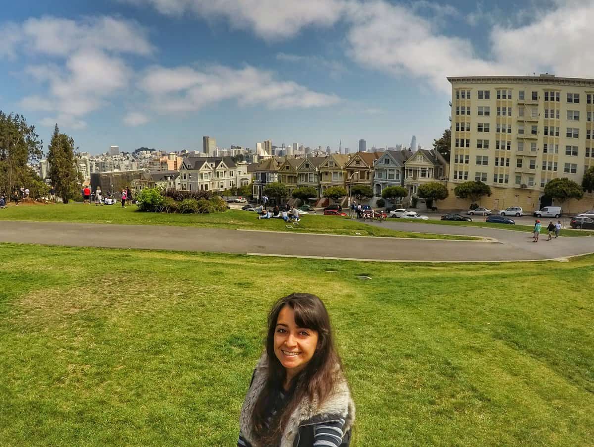 what to do in San Francisco in one day - Painted Ladies Victorian Houses A woman is standing and smiling in a park with a green grassy area in San Francisco, with the iconic Painted Ladies and city skyline in the background.