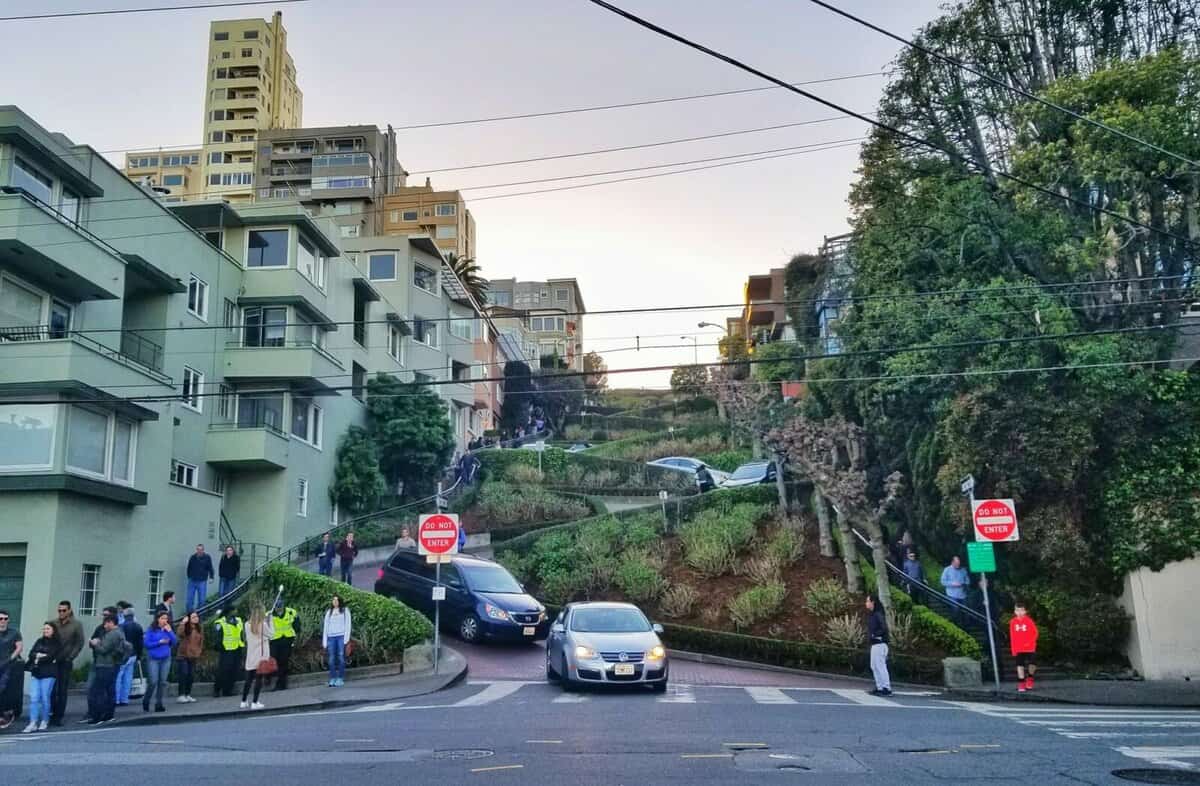 San Francisco in one day - Lombard Street A view of Lombard Street in San Francisco with cars navigating the famous winding road, surrounded by residential buildings and lush greenery, as people watch from the sidewalks.