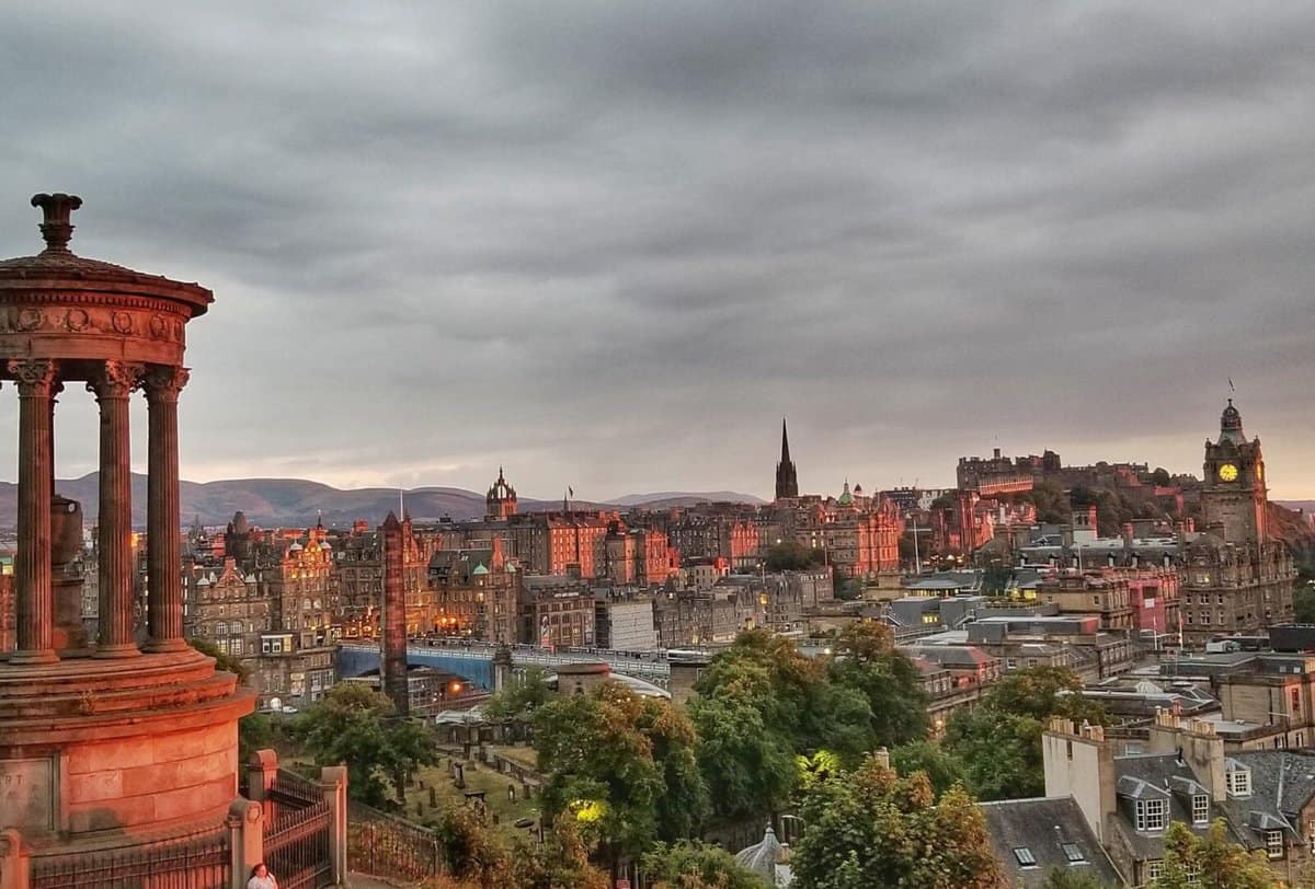 A scenic view of Edinburgh, Scotland, showcasing a historical monument in the foreground on the left and the city's old buildings, clock tower, and spires bathed in soft light, under a cloudy sky.