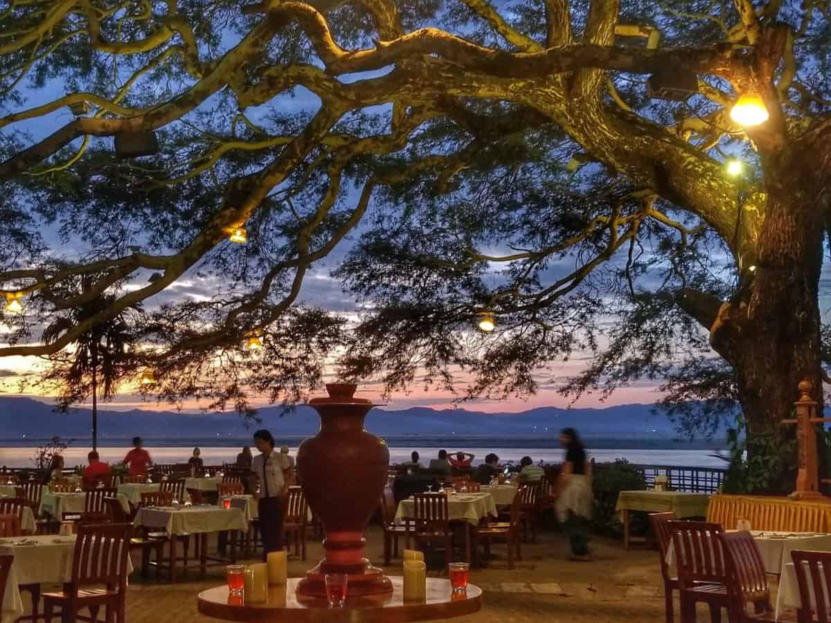 Outdoor dining area at Thande Hotel in Old Bagan, Myanmar, beautifully lit under a large tree during sunset with the river and mountains in the background.