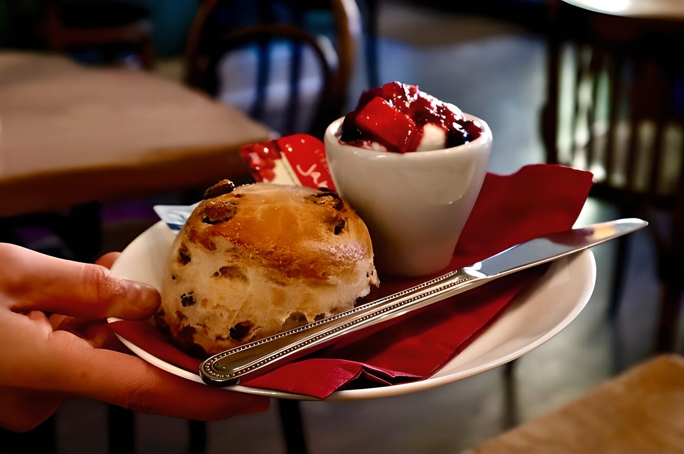 Close-up of a hand holding a plate with a traditional scone and a small bowl of cream topped with strawberry sauce in a café in Edinburgh, Scotland.