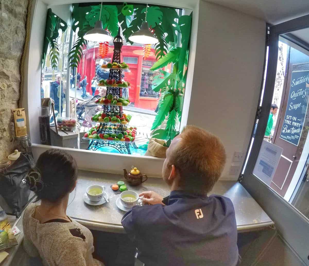 Two people seated at a table inside a cafe on Victoria Street in Edinburgh, Scotland, looking out of the window at a decorative display resembling the Eiffel Tower adorned with colorful fruits.