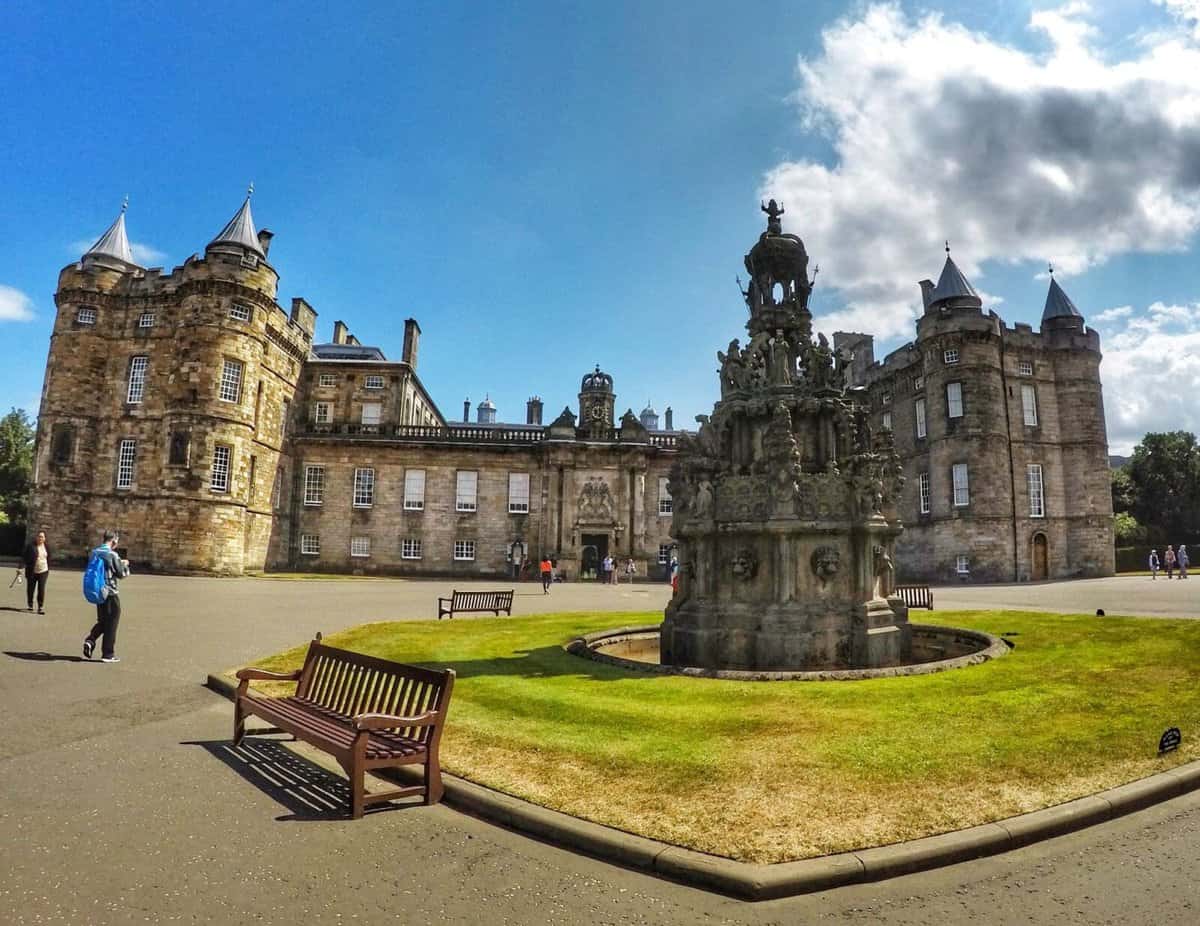 Holyrood Palace in Edinburgh, Scotland, with people walking in the foreground and a historic fountain structure in the center of the courtyard.