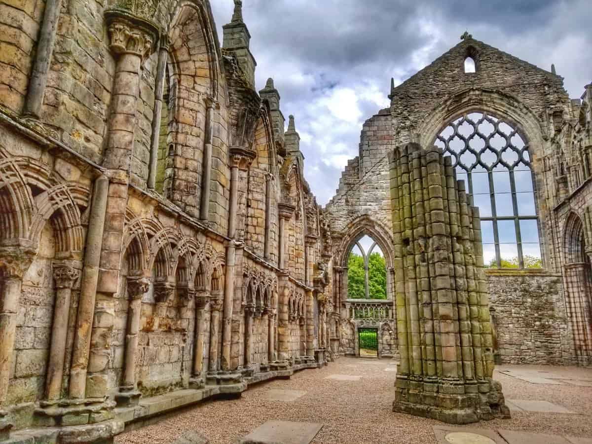Ruins of Holyrood Abbey in Edinburgh, Scotland, showcasing detailed stone craftsmanship with arches and columns, under a cloudy sky.
