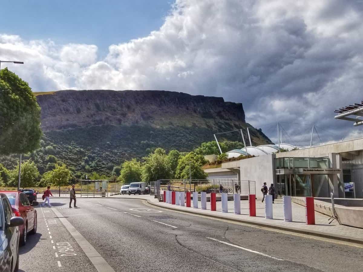 View of Arthur's Seat in Edinburgh, Scotland, seen from a nearby street with buildings, cars, and pedestrians.