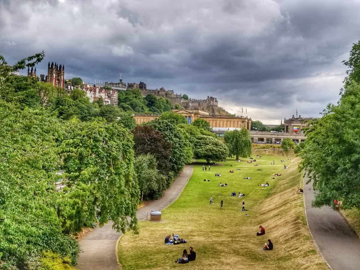 View of Princess Street Gardens in Edinburgh, Scotland, featuring lush greenery, scattered groups of people relaxing on the grass, historic buildings, and an overcast sky.