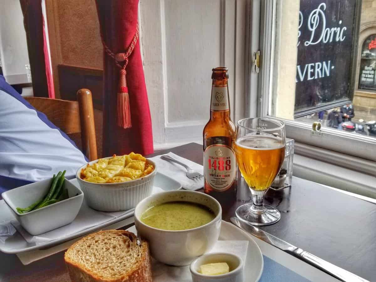 A table set for a meal at The Doric Tavern in Edinburgh, Scotland, featuring a bowl of soup with a slice of bread, a small bowl of green beans, a dish of macaroni pie, a bottle of beer, a glass of beer, and a small dish of butter, with a window displaying the tavern's sign in the background.