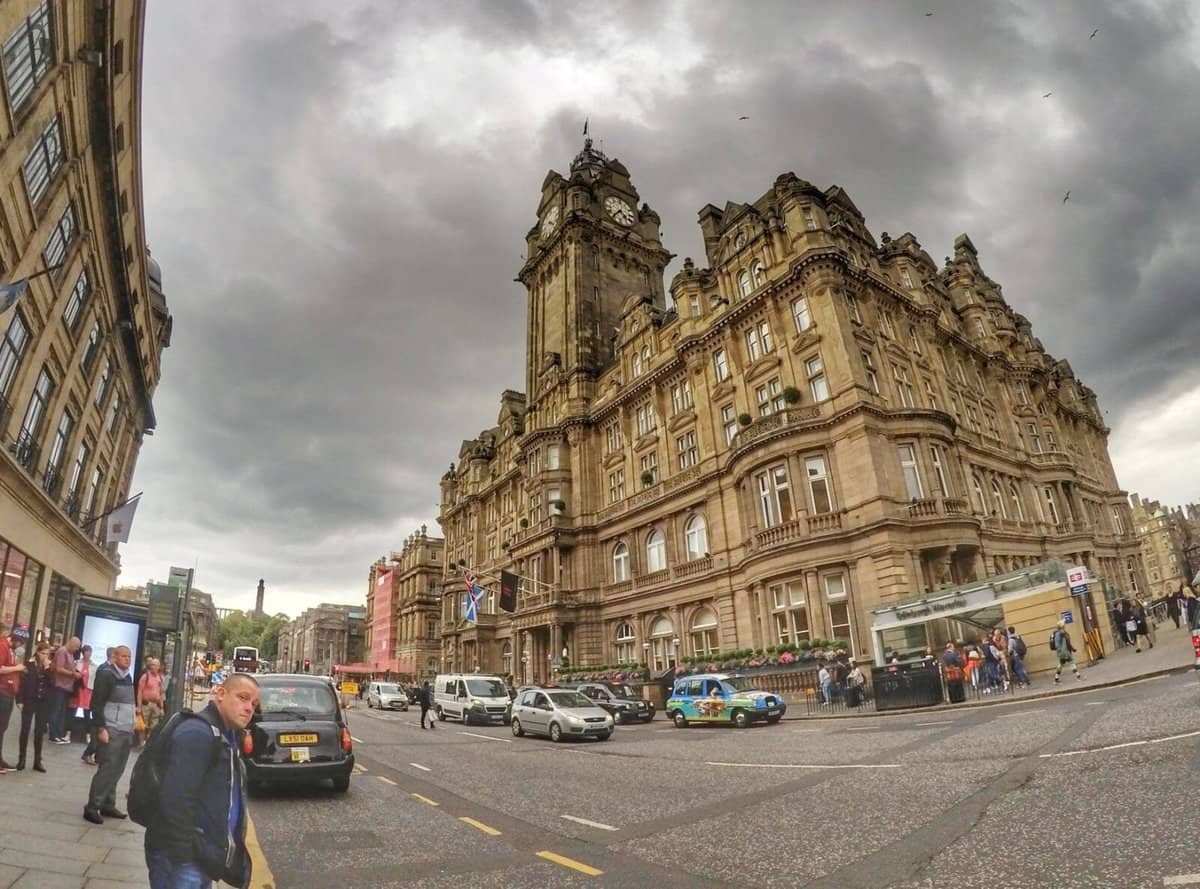 The Balmoral Hotel in Edinburgh, Scotland, with its iconic clock tower and grand Victorian architecture, seen against a cloudy sky. Pedestrians and vehicles occupy the bustling street in front.