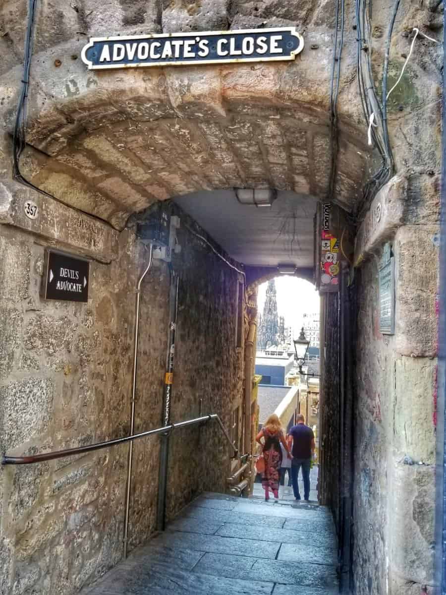 View of Advocate's Close on Royal Mile in Edinburgh, Scotland, showing a narrow cobblestone path between stone walls, with a sign reading 'Advocate's Close' at the top and several people walking down the path.