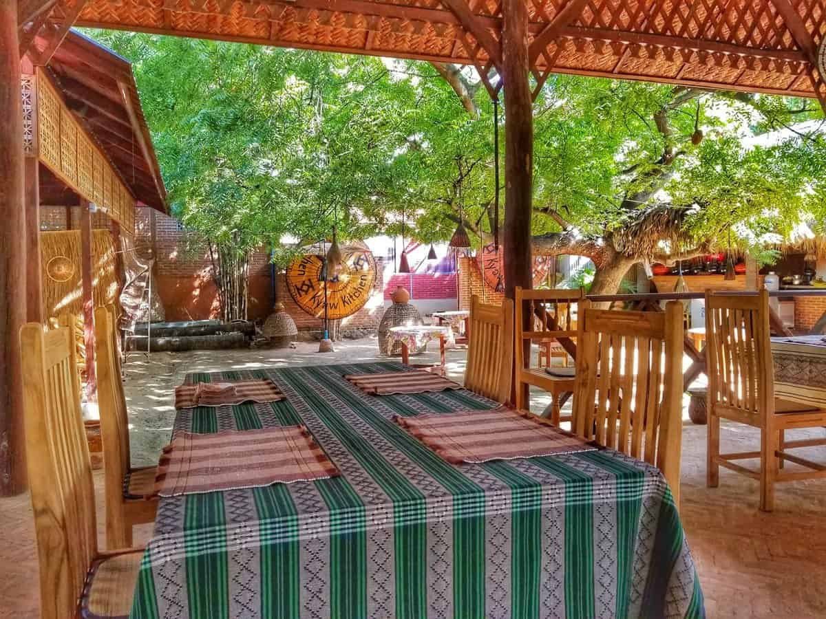 Outdoor dining area at a restaurant in New Bagan, Myanmar, with wooden tables and chairs, colorful tablecloths, and lush trees providing shade.