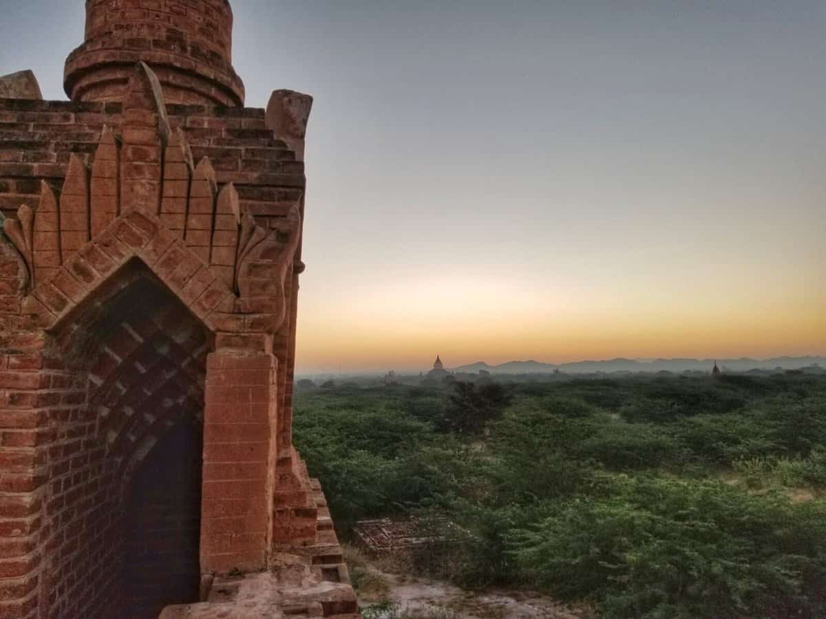 Sunset view of ancient temple ruins in Bagan, Myanmar, with lush vegetation and distant pagodas.