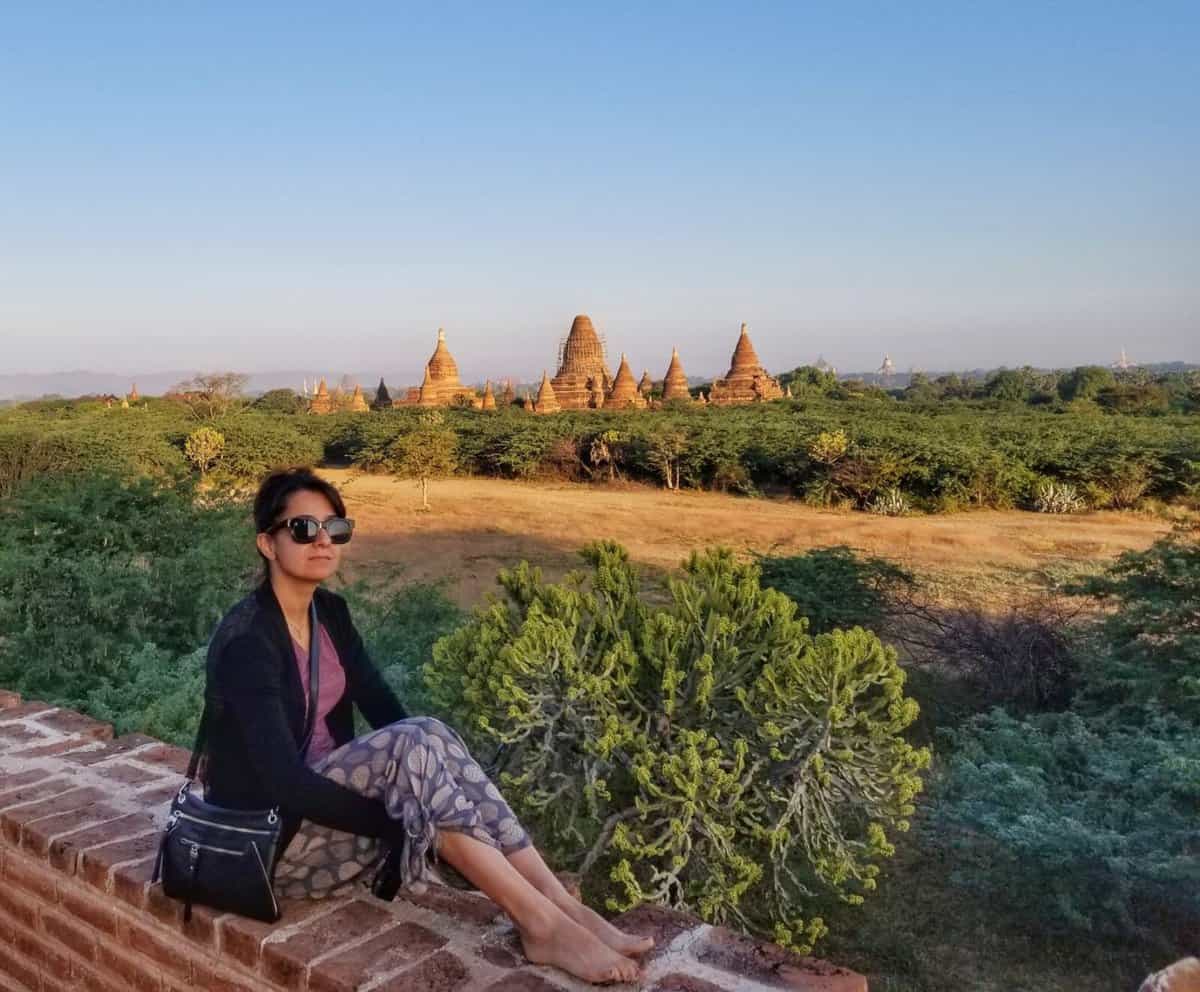 A person wearing sunglasses and casual clothing sits on a brick ledge, with a backdrop of ancient temples and green foliage in Bagan, Myanmar.