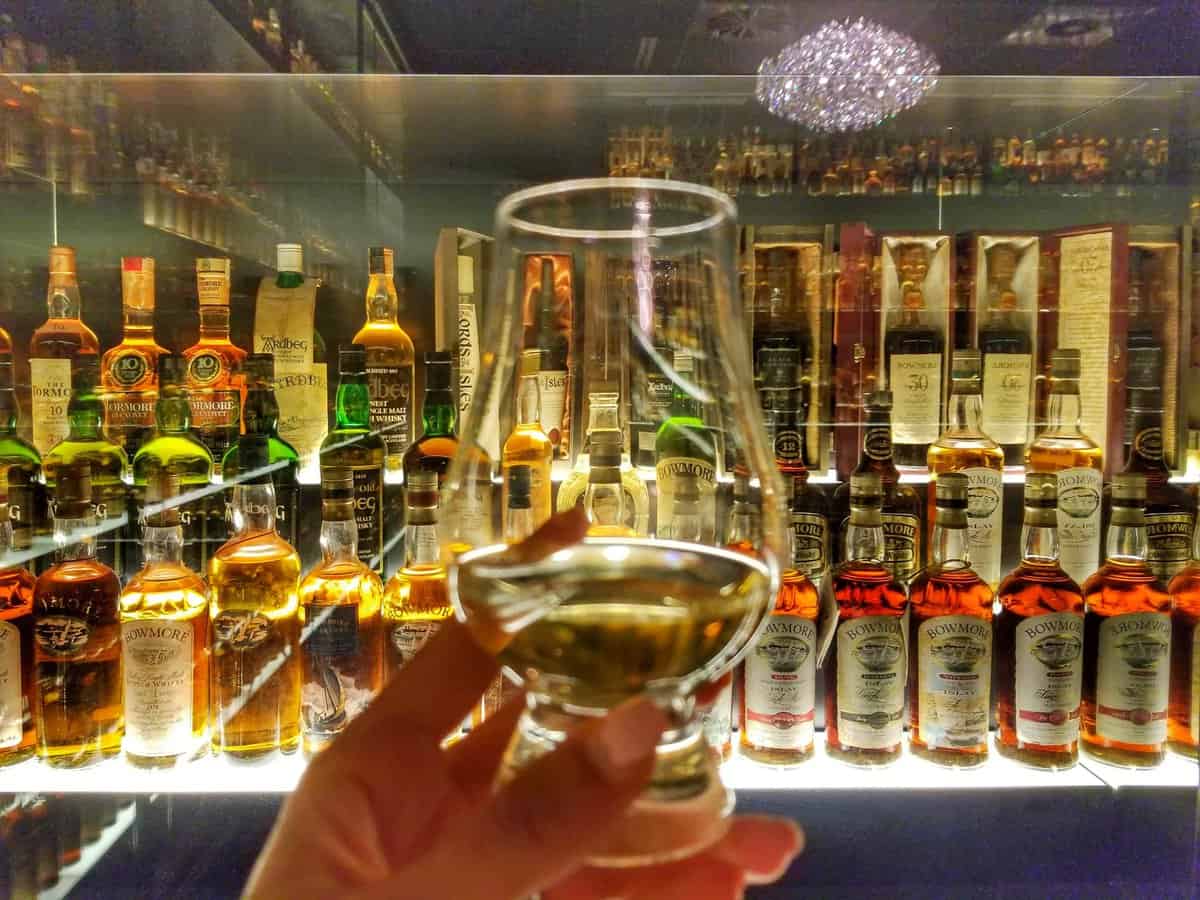 A display of various whisky bottles behind a glass, with a hand holding a filled whisky glass in the foreground at The Scotch Whisky Experience in Edinburgh, Scotland.
