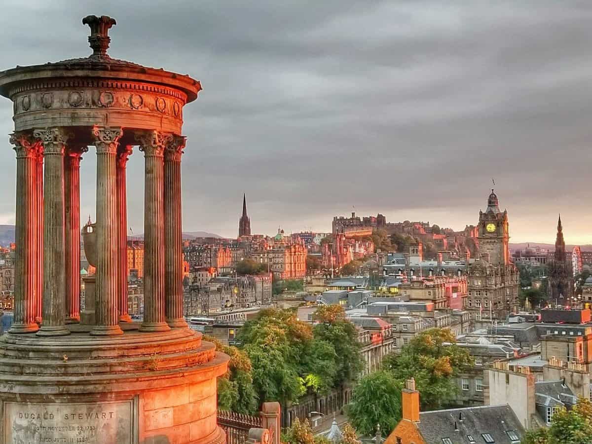 View from Calton Hill in Edinburgh, Scotland, showcasing the Dugald Stewart Monument in the foreground with Edinburgh's city skyline, including the Edinburgh Castle, in the background during sunset.