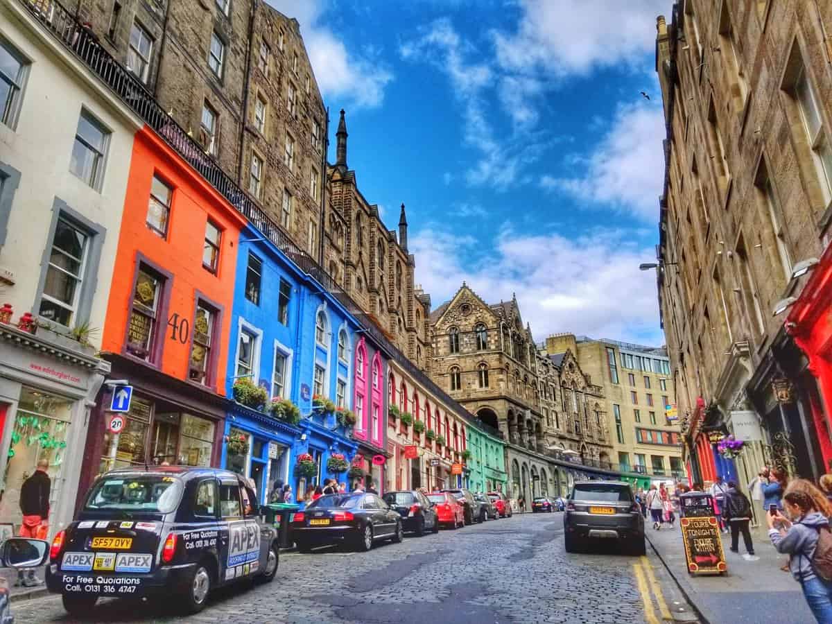 Colorful buildings along Victoria Street in Edinburgh, Scotland, with a mix of parked cars and pedestrians on a sunny day.