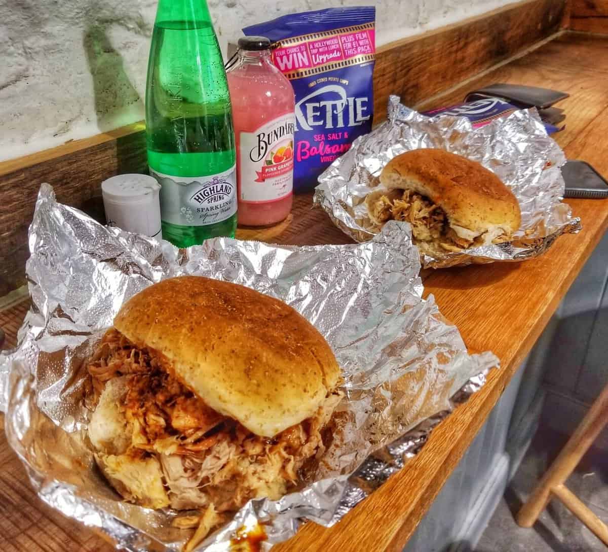 Two pulled pork sandwiches wrapped in foil on a wooden counter, accompanied by a bottle of sparkling water, a bottle of pink grapefruit soda, and a bag of Kettle Sea Salt & Balsamic Vinegar chips. The setting is an eatery on Victoria Street in Edinburgh, Scotland.