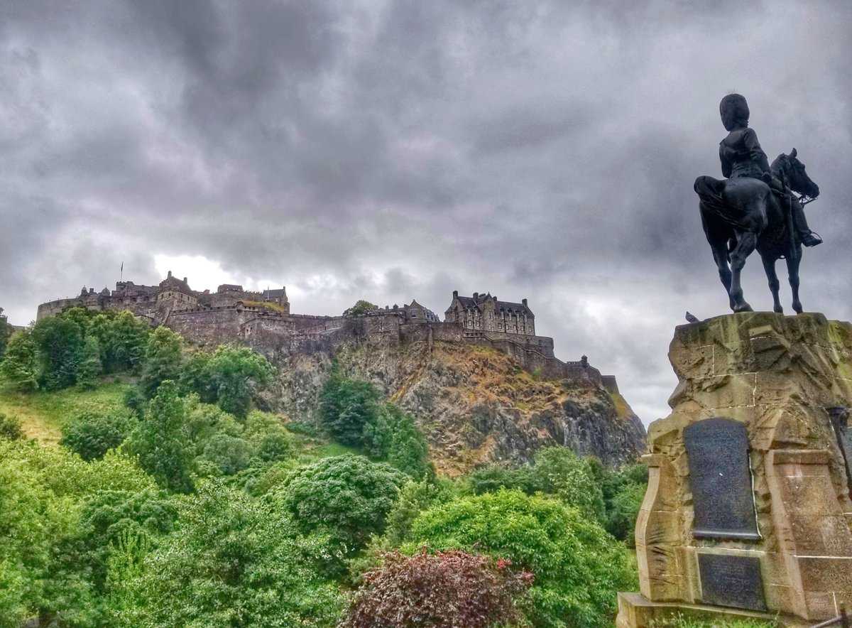 View of Edinburgh Castle perched on a rocky hill with lush greenery in the foreground and a statue of a rider on horseback in Edinburgh, Scotland under a cloudy sky.