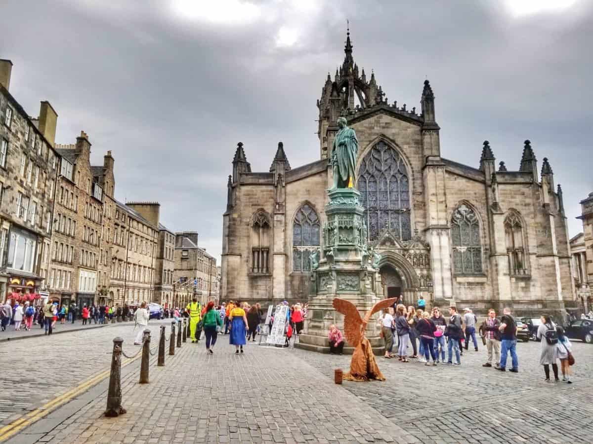 A bustling scene on the Royal Mile in Edinburgh, Scotland, with crowds of people walking along the cobblestone street. In the background stands the historic St Giles' Cathedral with its gothic architecture. A statue and a street performer in a winged costume can be seen in the foreground.