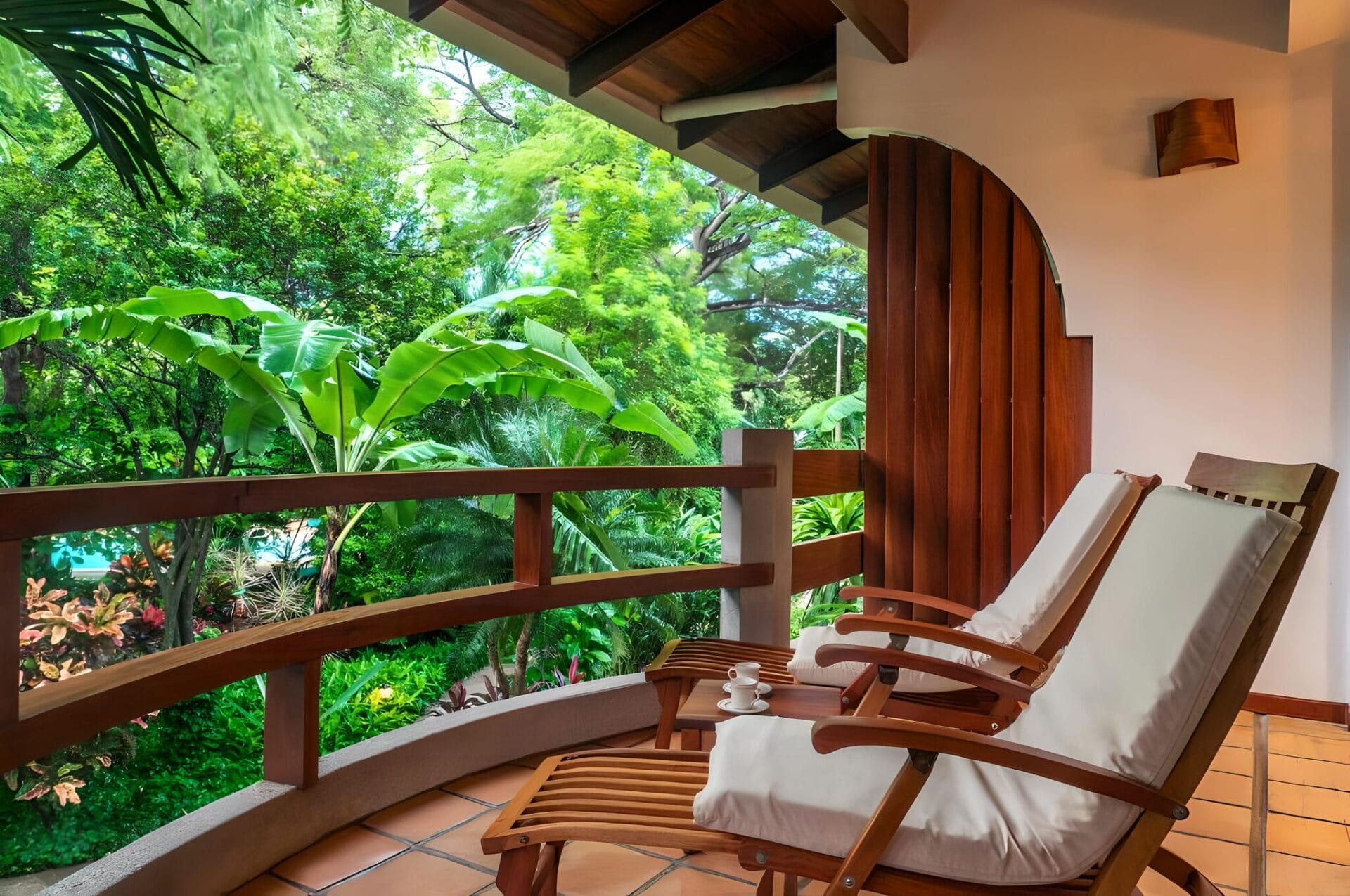 Two wooden lounge chairs with white cushions on a balcony surrounded by lush greenery at Capitán Suizo Beachfront Boutique Hotel in Tamarindo, Costa Rica.