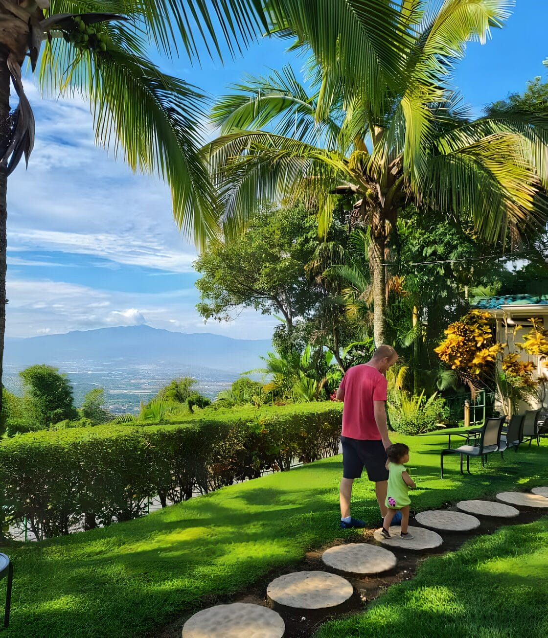 Alajuela Buena Vista Chic Hotel Costa Rica last day A man and child walking on a stone path surrounded by lush green grass and tropical plants at Buena Vista Chic Hotel in Alajuela, Costa Rica, with a scenic mountain view in the background.