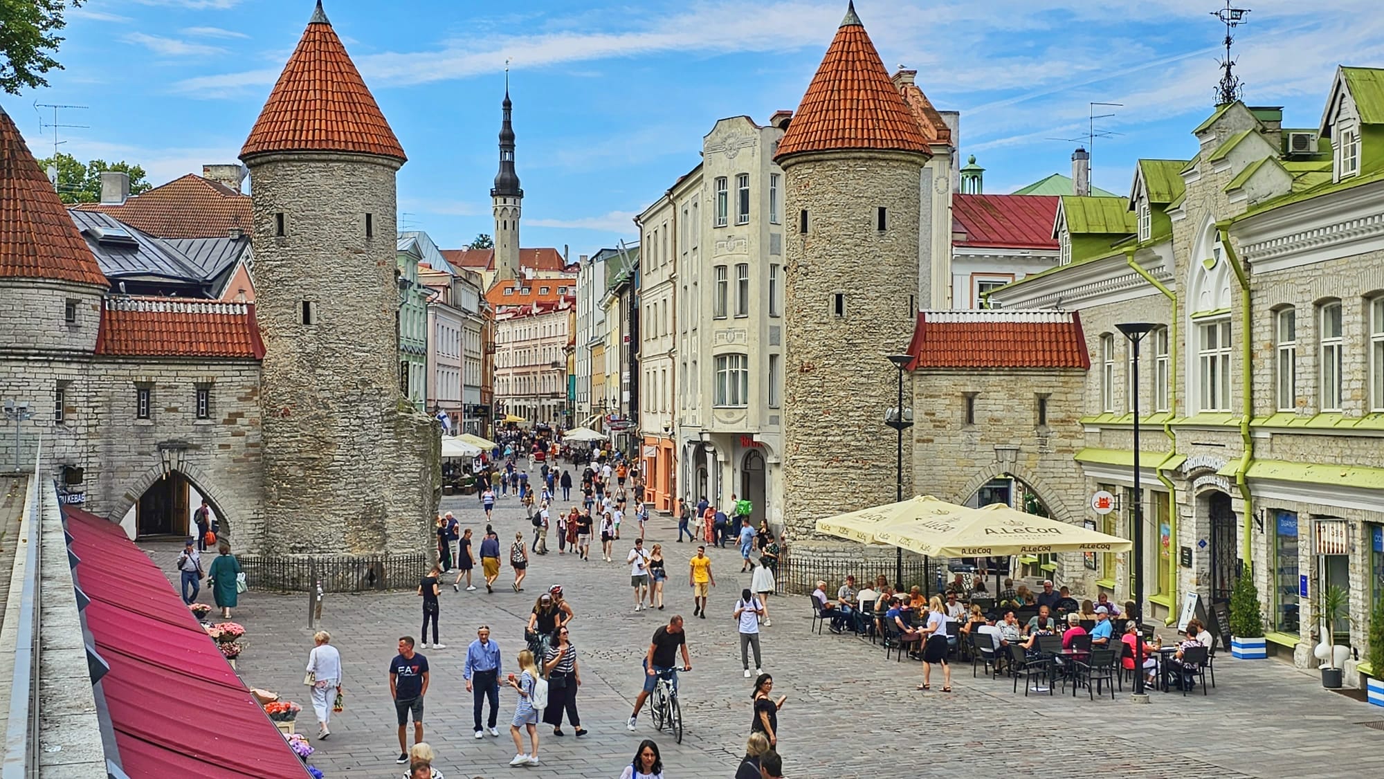 A bustling scene at the Old Town Gate in Tallinn, Estonia, showcasing historic stone buildings with red conical roofs, a lively street lined with people, and outdoor cafes.