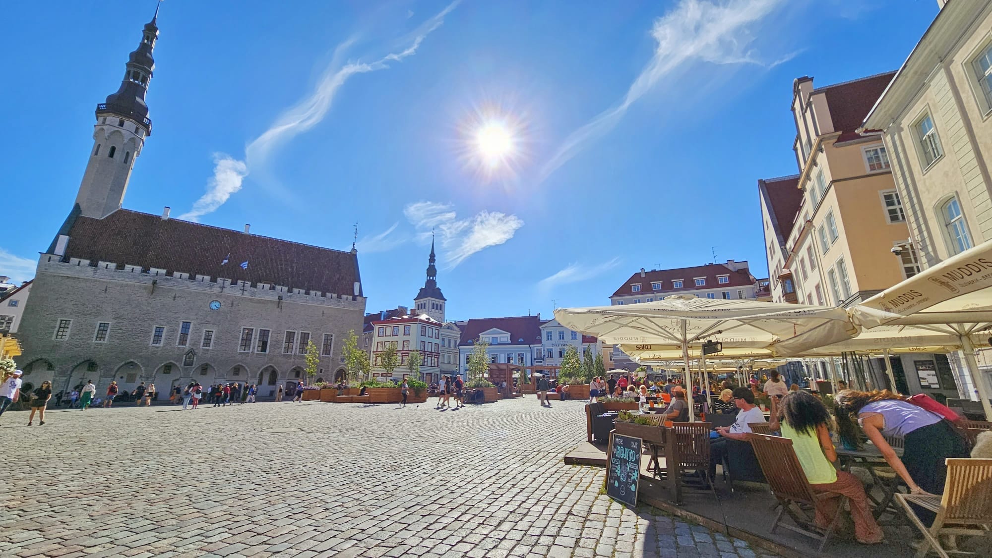 Town Hall Square in Tallinn, Estonia under a bright sunny sky with people sitting at outdoor cafes and strolling in the square.