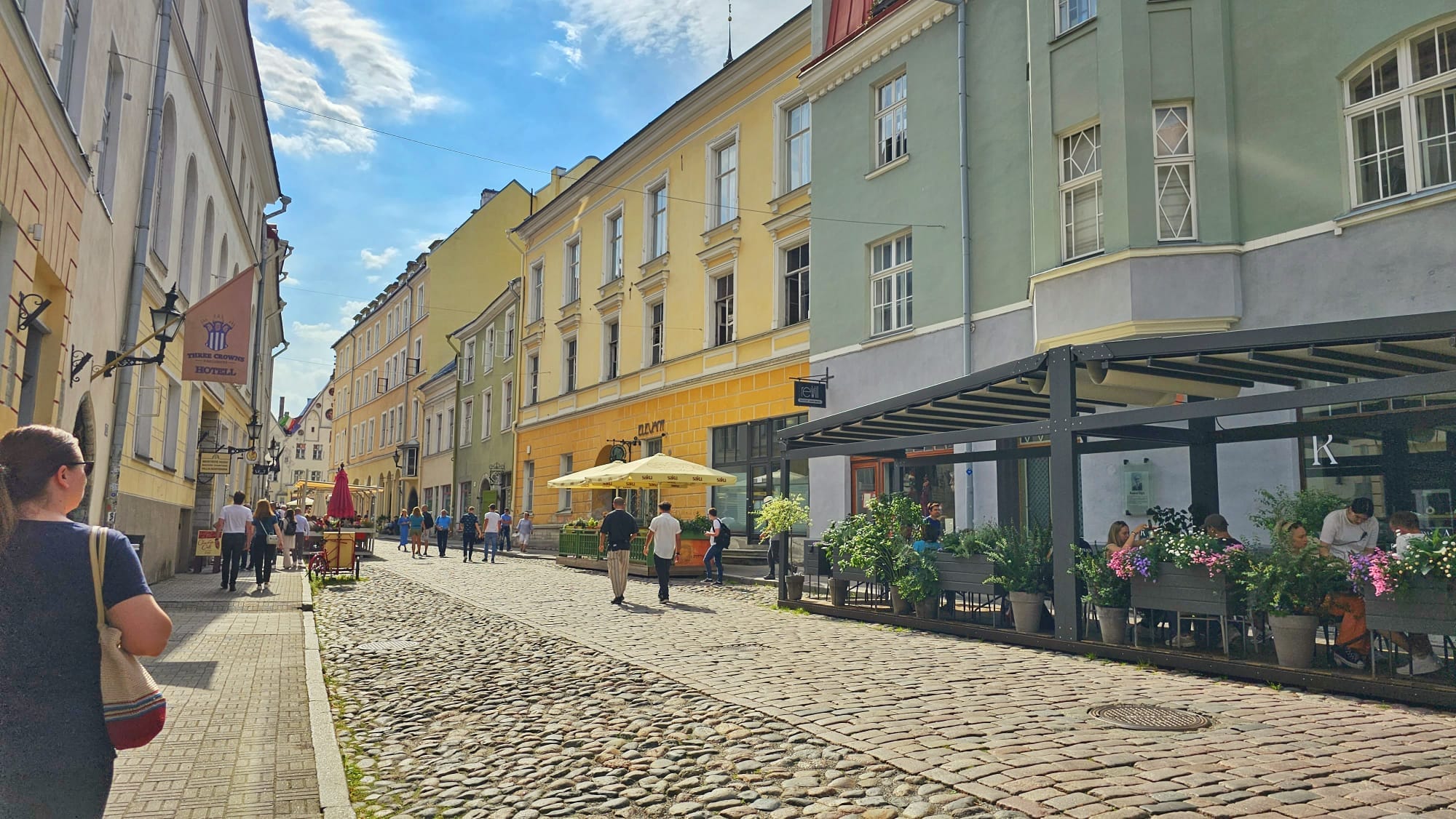 A cobblestone street lined with colorful buildings and outdoor cafes in Tallinn, Estonia. People are walking along the street, enjoying a sunny day.