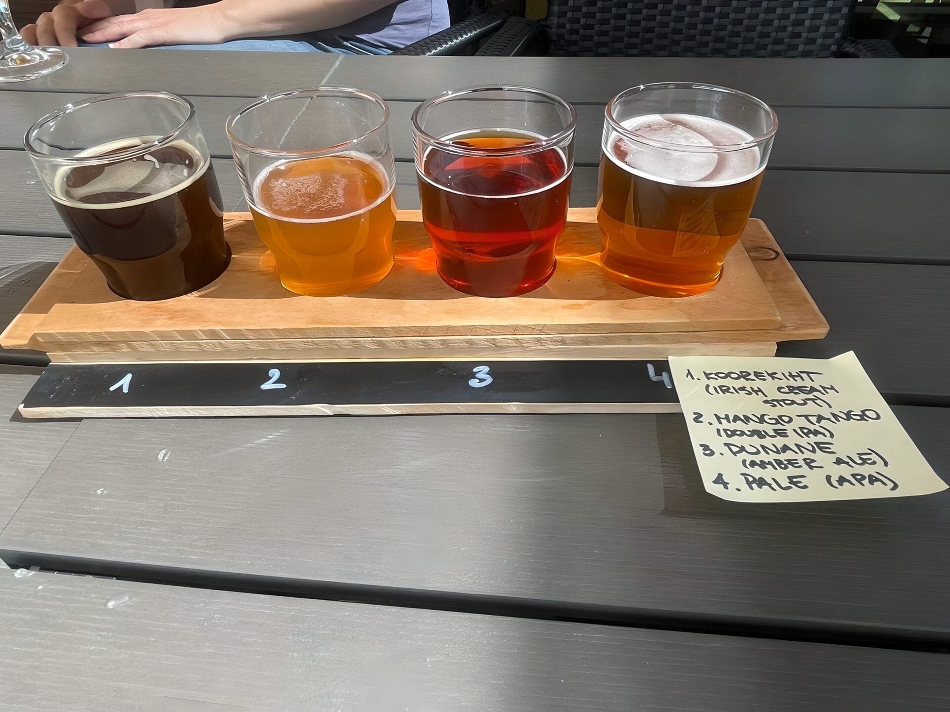 A wooden flight board with four glasses of different types of beer, each glass is labeled with numbers 1 through 4. A handwritten note beside the flight board lists the beers: 1. Koorekinit (Irish Cream Stout), 2. Mango Tango (Double IPA), 3. Punane (Amber Ale), and 4. Pale (APA). The setup is on an outdoor table in Tallinn, Estonia.