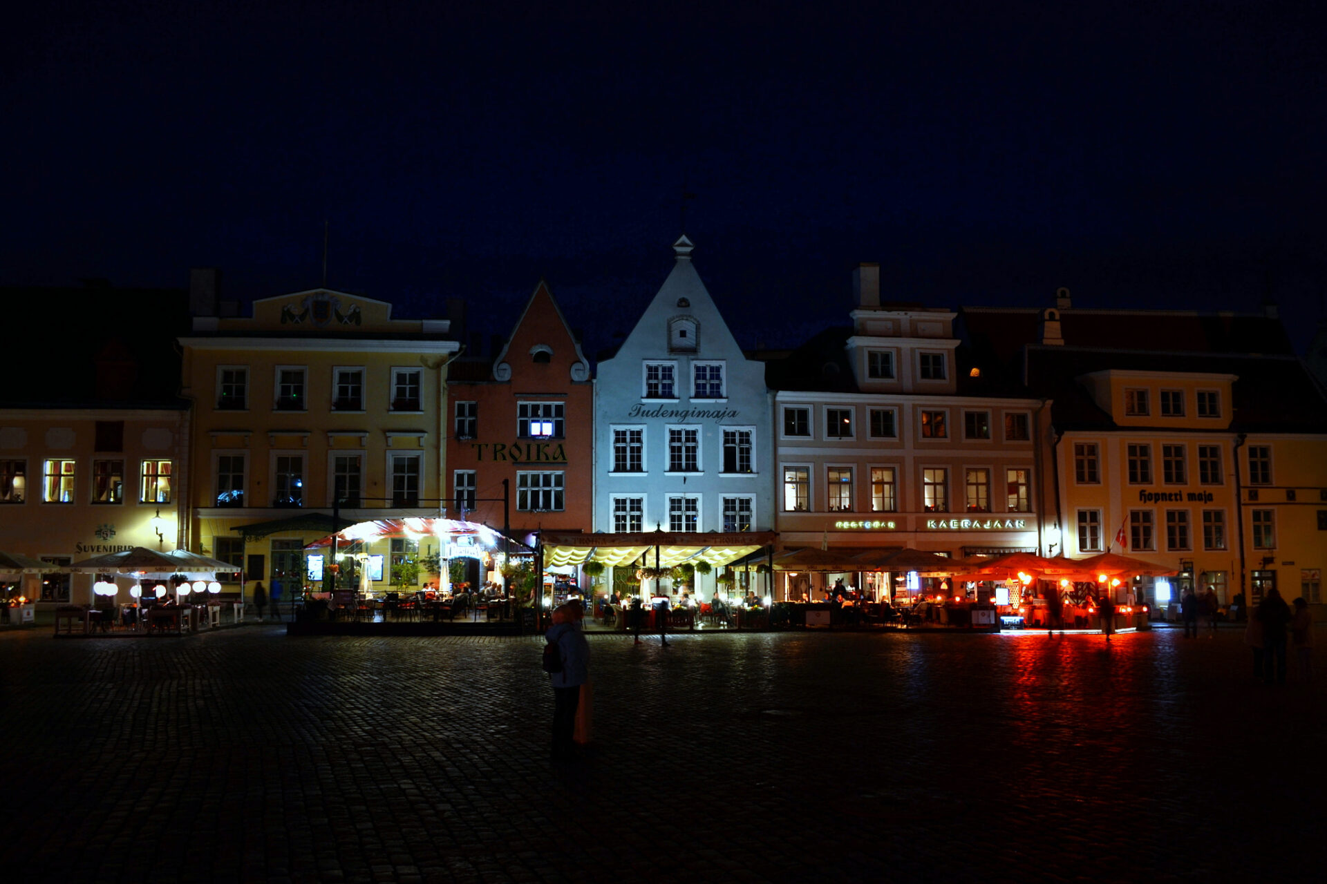 Night view of a bustling town square in Tallinn, Estonia, with illuminated buildings and outdoor cafes.