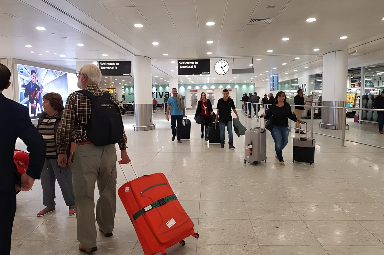 Travelers walking through the terminal at Edinburgh Airport in Edinburgh, Scotland. Some are carrying luggage, while others are walking with rolling suitcases. The terminal is well-lit, with signs indicating arrivals and directions.