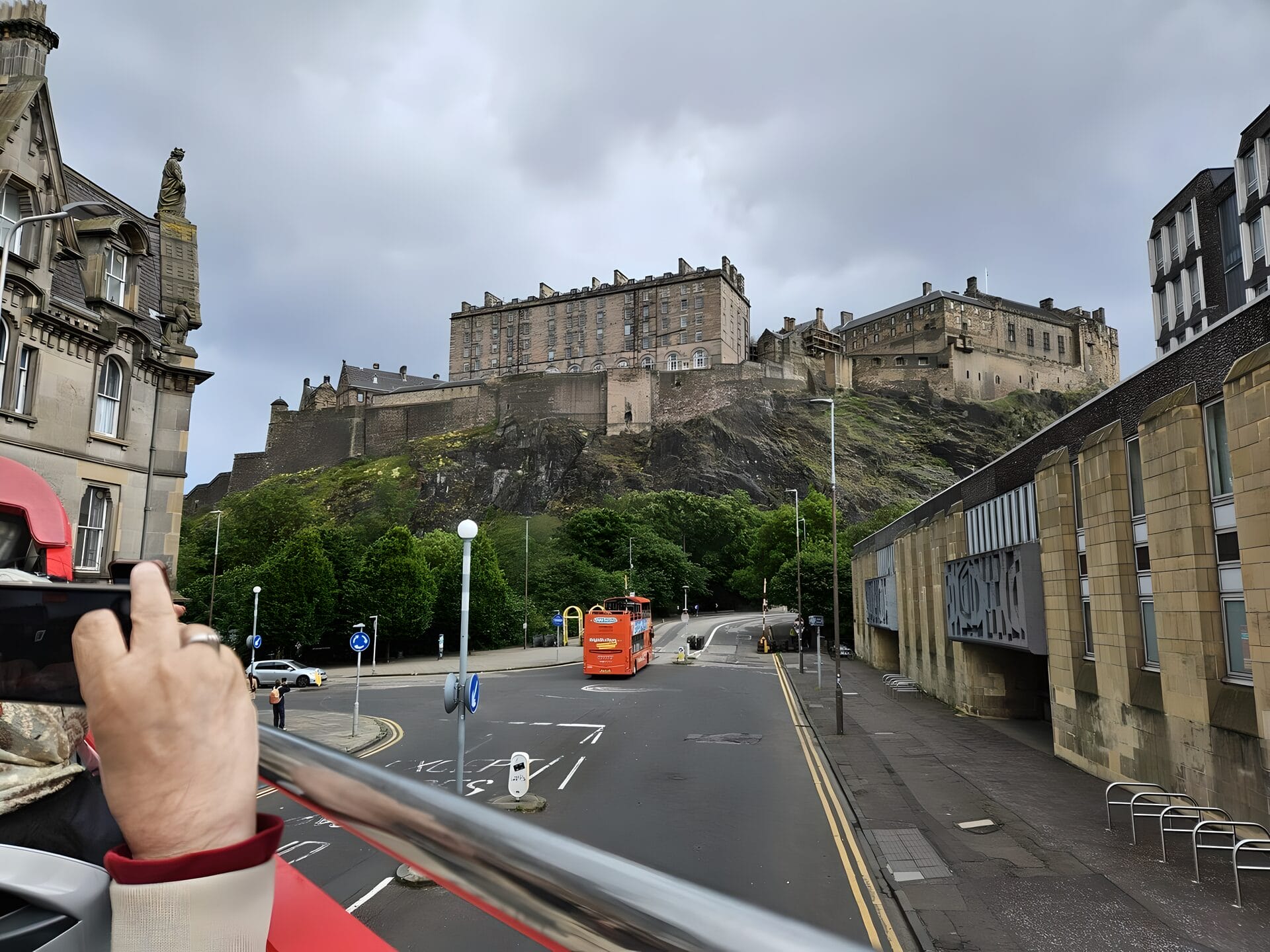 A view of Edinburgh Castle taken from a Hop-On Hop-Off bus in Edinburgh, Scotland. The castle appears elevated on a rocky hilltop, with classic architectural buildings, greenery, and an overcast sky in the background. A person on the bus is capturing the scene with their phone.