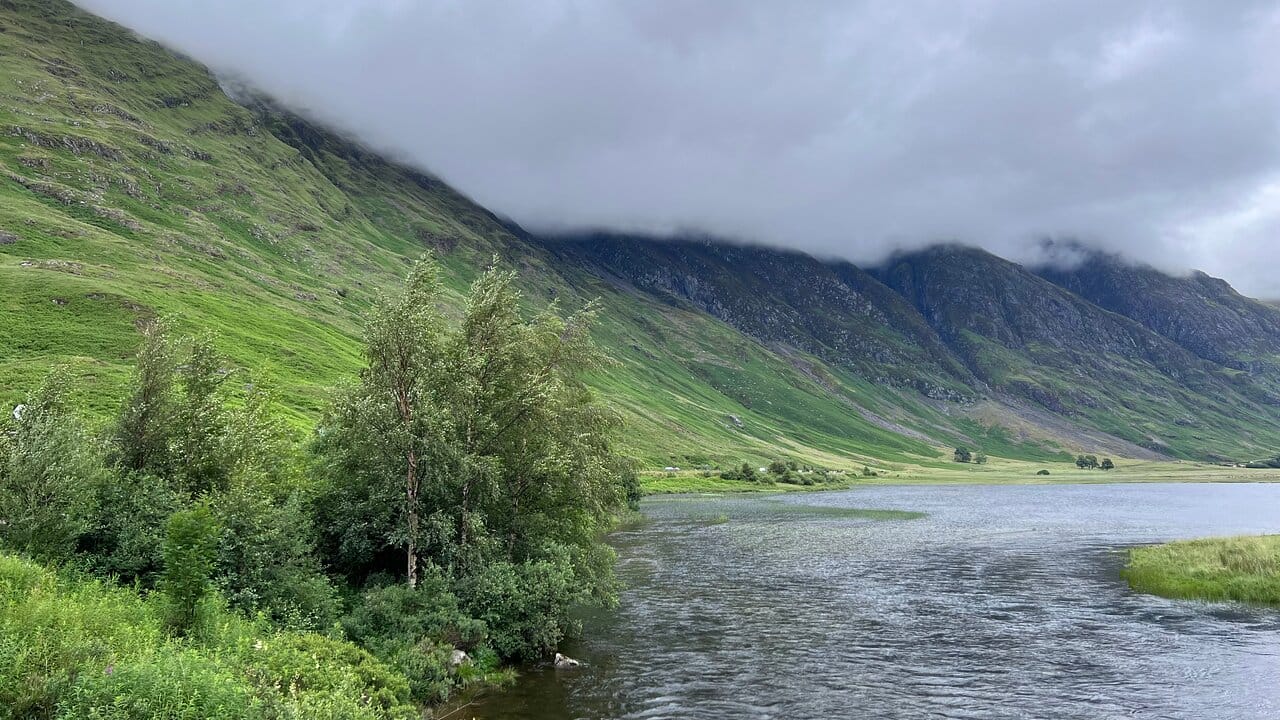 Scenic view of the Scottish Highlands with low-hanging clouds over misty green hills and a tranquil river in the foreground.
